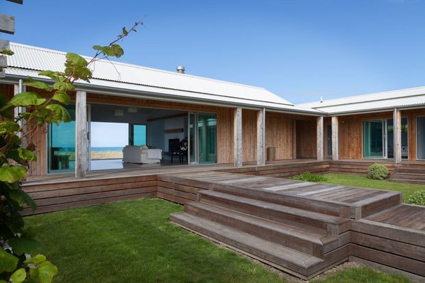 Single-storey coastal home arranged around a landscaped courtyard, with weathered timber cladding and decking, broad glazed openings, and a low corrugated metal roof. A stepped timber deck leads through a grass courtyard toward the living areas, while the ocean is visible through the open glazing in the background.