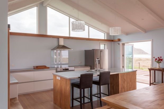Bright open-plan kitchen and dining area with a large timber-fronted island, pale stone benchtop, integrated cabinetry, stainless-steel appliances and three dark upholstered bar stools. The room has expansive angled glazing, a pitched ceiling, timber flooring and warm timber detailing, with a glazed opening to an outdoor deck and coastal landscape beyond.
