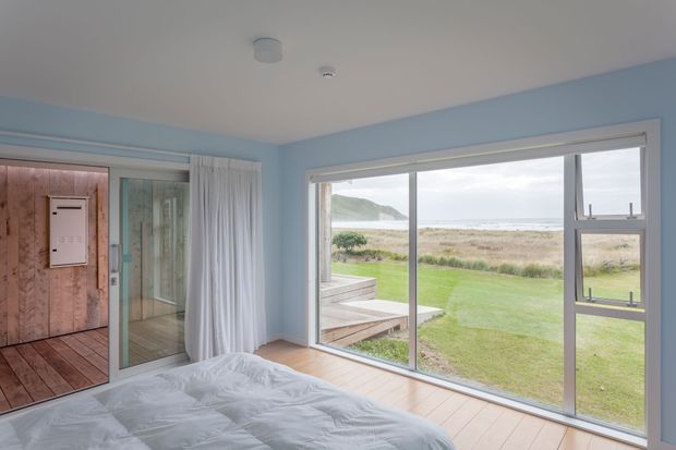 Bright coastal bedroom with pale blue walls, light timber flooring, and a large sliding glass window/door opening to a lawn, dunes, and ocean beyond. The room includes a bed, white curtains, and a timber-clad adjoining entry or bathroom area visible at left.