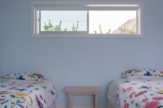 A compact twin bedroom with two single beds positioned beneath a horizontal window. The room has pale blue walls, white trim, a simple central timber bedside table, and bright floral bedding. Natural light enters through the partially shaded window, creating a simple, cheerful shared sleeping space.