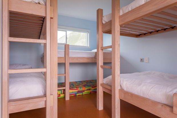 A compact shared bunk room with multiple built-in or freestanding timber bunk beds, white bedding, pale blue walls, a small window, and a colourful patterned rug on a warm brown floor. The room is practical, bright, and designed for children or guests.