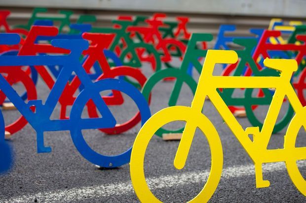 A close-up outdoor view of numerous brightly painted metal bicycle-shaped barriers or bike racks arranged across a paved surface, with prominent yellow bicycles in the foreground and red, blue, and green bicycles behind them. The image is colorful and graphic, with shallow depth of field and no visible interior architecture.