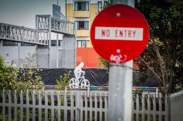 Urban streetscape with a prominent red 'No Entry' sign in the foreground, metal pedestrian barriers and fencing, construction hoarding, and a multi-storey building with industrial-style elevated walkways and colourful facade elements in the background.