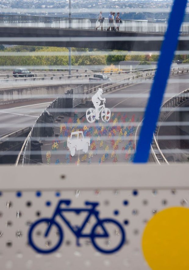 View through a glazed or reflective structure toward a multi-level urban transport interchange, with elevated pedestrian walkways, road lanes, colorful bicycle artwork, and a large blue bicycle graphic in the foreground. A distant built-up hillside is visible beyond the infrastructure.