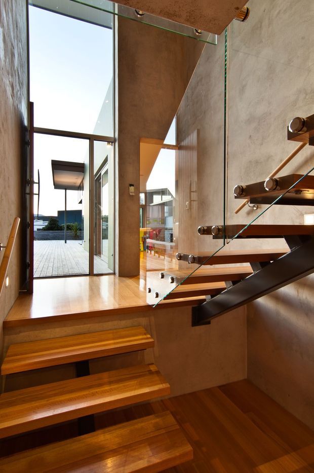 Interior view of a modern stairway with floating timber treads, dark steel supports, and a clear glass balustrade. Warm timber flooring and stair surfaces contrast with textured concrete or plaster walls. Full-height glazing opens toward a bright outdoor terrace, while a living area is visible beyond the landing.