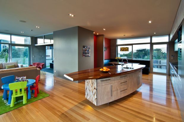 Open-plan contemporary kitchen and living area with a large curved island featuring a polished dark timber benchtop, pale timber cabinetry, and a textured stone-clad end panel. The room has extensive floor-to-ceiling glazing, warm timber flooring, recessed ceiling lights, and a connected lounge and children's play area. A balcony or exterior view is visible through the windows.