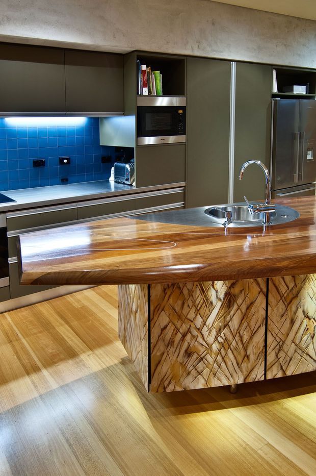 A sophisticated modern kitchen featuring dark flat-panel cabinetry, integrated appliances, a vivid blue tiled splashback, and a large polished timber island with a curved overhang and integrated stainless-steel sink. The island front has distinctive sculptural textured timber panels, complemented by warm timber flooring and brushed metal accents.