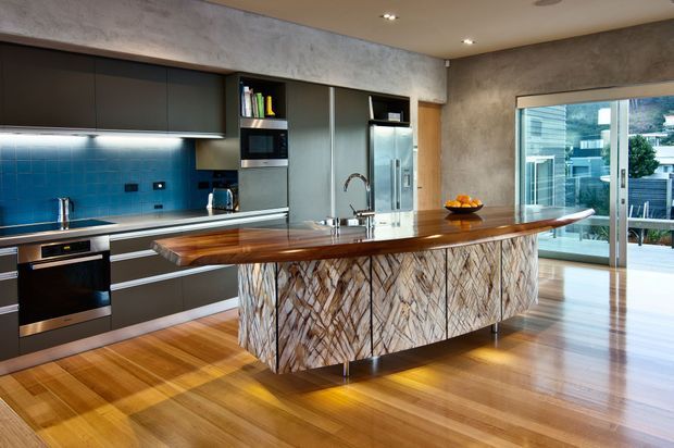 Contemporary kitchen with dark flat-panel cabinetry, integrated stainless-steel appliances, a vivid blue tiled splashback, and a large sculptural island featuring a thick timber benchtop and patterned stone-look cabinetry. Warm timber flooring, concrete-look walls, under-cabinet lighting, and glazed doors opening to an outdoor terrace create a polished, open and inviting space.