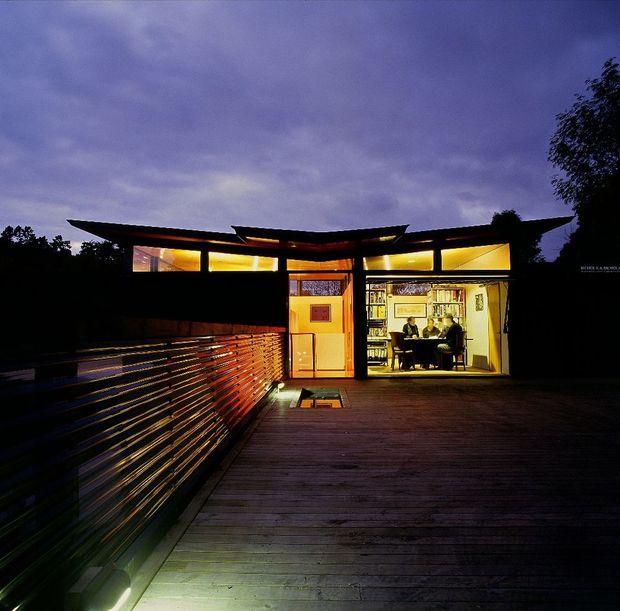 Twilight exterior view of a compact modern house or pavilion opening onto a broad timber deck. The building has a low, angular roofline, extensive warm-lit glazing, and a horizontal slatted timber privacy screen or balustrade along the deck. Through the large opening, a warmly illuminated interior with shelving, artwork, and a small table is visible. The atmosphere is dramatic and moody, with dark surrounding vegetation and a purple evening sky.