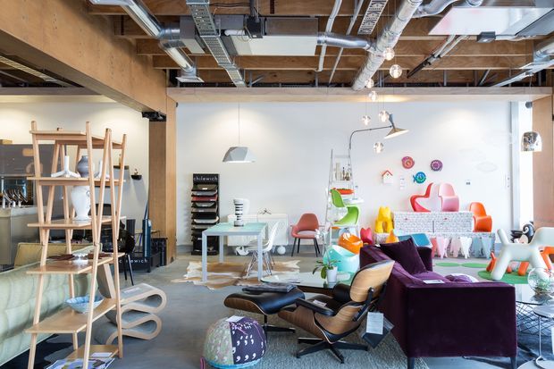 Bright open-plan commercial interior with a creative studio or showroom atmosphere. The space features exposed timber ceiling structure, extensive exposed ductwork and services, polished concrete flooring, white walls, suspended pendant lighting, display shelving, worktables, colourful contemporary furniture, and a prominent mid-century-style lounge chair with ottoman. The room combines an office workspace with a furniture and product display area.