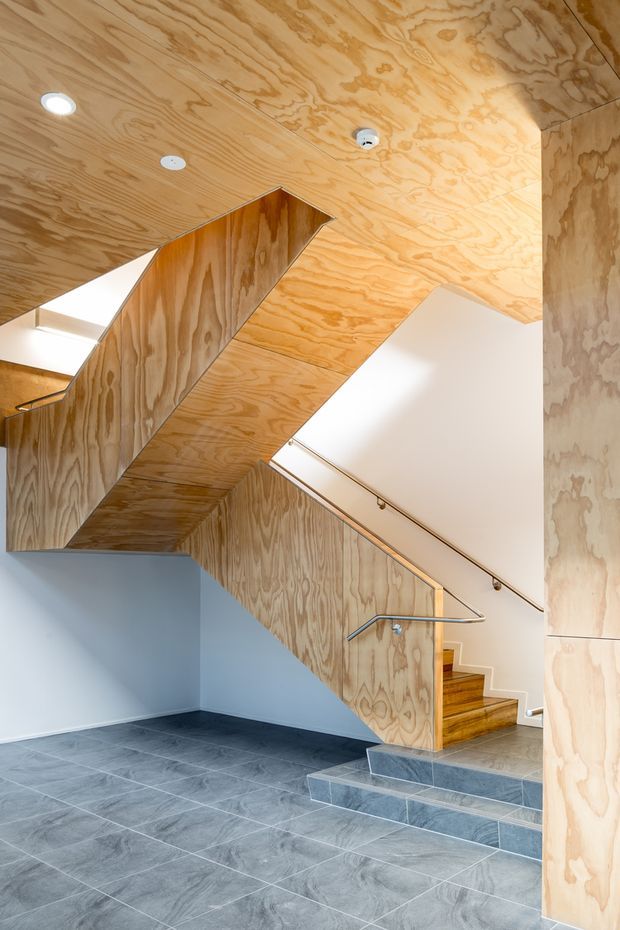 Interior view of a contemporary stairway with plywood-clad walls, ceiling, balustrade and stair surfaces. The stair rises from a grey tiled floor, with a simple metal handrail, recessed ceiling lights and white plasterboard walls creating a bright, clean circulation space.