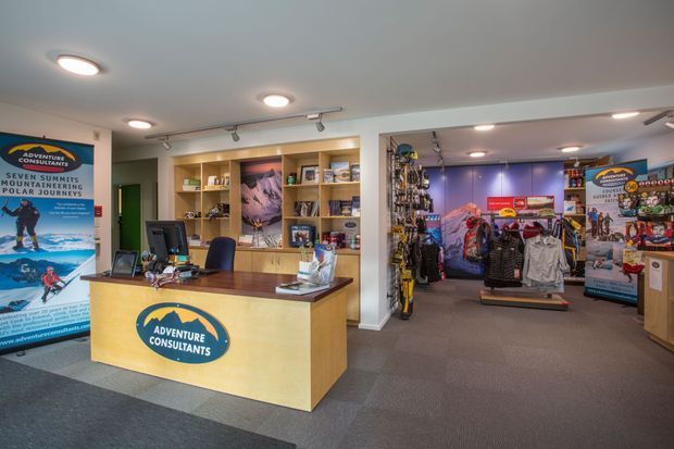 A bright commercial adventure and outdoor equipment retail space with a timber reception counter in the foreground, branded signage, built-in timber display shelving, computers, and a retail area displaying outdoor clothing and gear beyond. Recessed and track lighting illuminate grey carpeted floors and neutral walls.