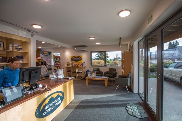 A bright commercial reception and waiting area for Adventure Consultants, with a timber reception counter and branded signage in the foreground, open shelving and promotional displays along the left wall, and a small seating area with sofas and armchairs by a large window. Full-height glazed entry doors on the right provide views to the street and neighbouring buildings. The space has a neutral palette, recessed ceiling lights, carpet flooring, and a practical contemporary fit-out.