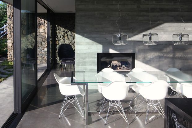 A contemporary dining area with a long glass dining table and multiple white molded-shell chairs on wire bases. Full-height black-framed glazing opens toward a stone exterior wall, while the interior features a dark concrete-look feature wall, a linear fireplace, and three suspended glass pendant lights. Strong sunlight creates geometric shadows across the room.