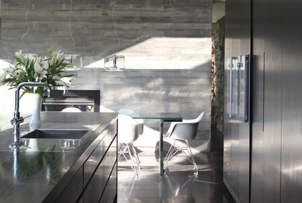 A sunlit contemporary kitchen with a long dark island, integrated stainless-steel sink and tall curved faucet, dark built-in cabinetry, and a compact dining area with white molded chairs around a glass-topped table. The room features a horizontally textured grey feature wall, polished dark flooring, and strong natural light creating a calm, refined atmosphere.