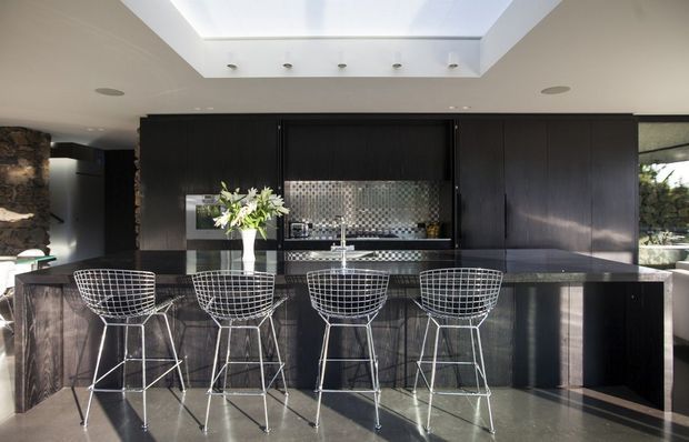 A dramatic contemporary kitchen with a large dark island and four wire-frame bar stools. Full-height black cabinetry spans the rear wall, incorporating a metallic mosaic tile splashback, sink, tapware and integrated appliances. A large roof skylight provides strong natural light, while stone detailing and glazing appear at the edges of the room.
