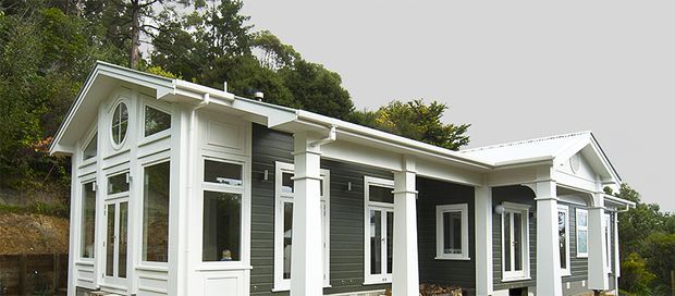 A single-storey white-and-charcoal residential house set against a wooded hillside. The home has a low-pitched gabled roof, dark horizontal weatherboard cladding, prominent white trim, tall multi-pane windows, classical porch-like columns, and decorative gable detailing. A large round feature window is visible in the front gable.