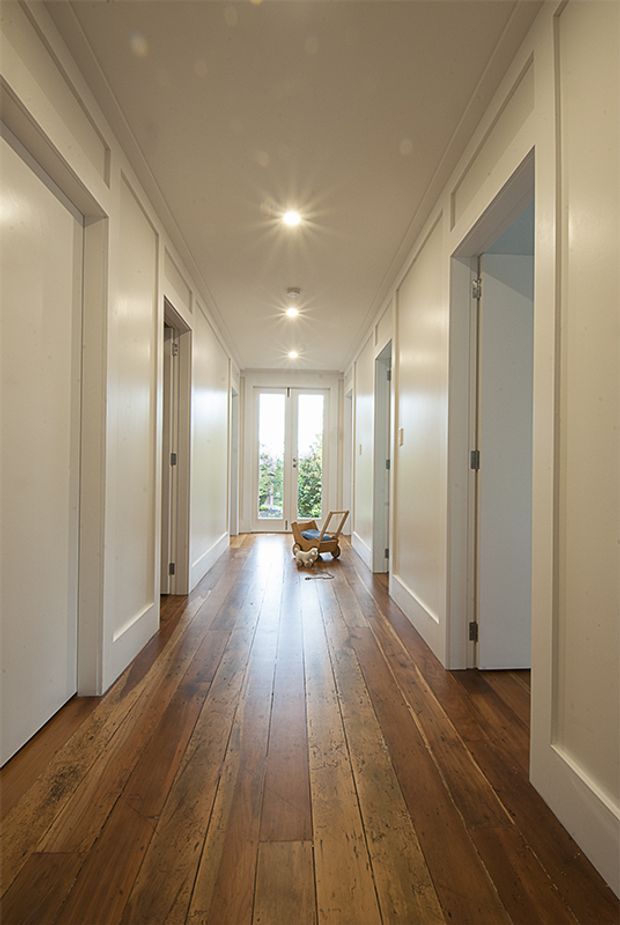 A long, bright residential hallway with white paneled walls, white door and window trim, recessed ceiling spotlights, and wide-plank warm timber flooring. Multiple rooms open off both sides, while glazed doors at the far end bring in natural light. A small wooden chair and decorative object sit near the end of the corridor.