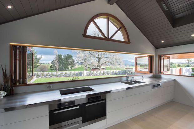A bright contemporary kitchen with a long white built-in cabinetry run, pale stone benchtops, integrated black appliances and a double sink beneath expansive picture windows. Timber-framed folding or sliding windows open the room to a landscaped rural outlook, while a high pitched ceiling with dark timber lining and a distinctive arched clerestory window adds architectural character.
