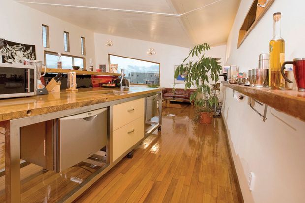 A warm, open-plan kitchen with extensive timber cabinetry and benchtops, stainless-steel appliances, and polished timber flooring. The room includes a central island with a sink, a raised red-accented breakfast or work surface, open shelving, and a small living area visible beyond. Tall narrow windows and large glazing provide natural light, while a potted indoor tree adds greenery.