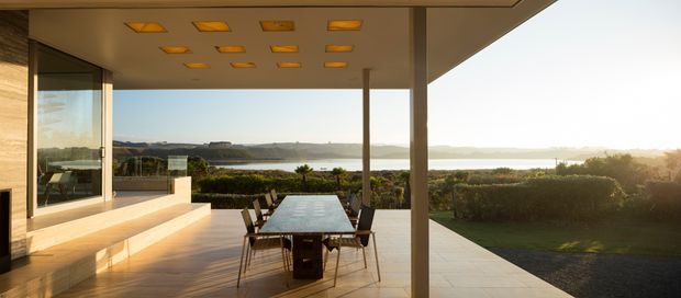 A refined outdoor dining terrace beneath a broad flat-roofed canopy, overlooking landscaped grounds and a calm body of water. The space includes a long dining table with chairs, built-in stepped seating or platforms, expansive glazing, warm timber-toned surfaces, and recessed amber-toned ceiling lights. The scene has a clean contemporary architectural character with strong indoor-outdoor connection and expansive waterfront views.