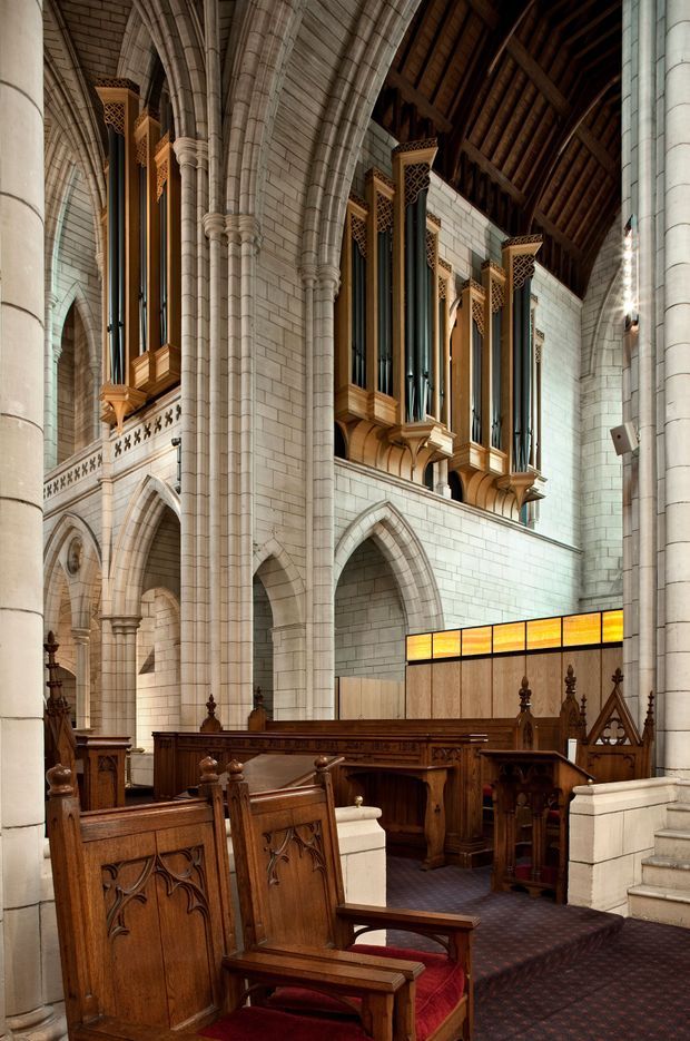 Interior view of a grand Gothic Revival church or cathedral, showing soaring pointed stone arches, clustered columns, ribbed vaulted construction, a dark timber roof, and a prominent set of tall pipe-organ pipes in carved timber cases. The foreground contains dark carved timber choir stalls and ecclesiastical seating with red upholstery, while a warm amber stained-glass-like partition adds colour at the rear.