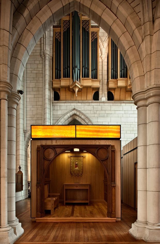 Interior view of a grand Gothic Revival church or cathedral, framed by large pointed stone arches and clustered columns. A prominent pipe organ with dark vertical pipes and warm timber detailing occupies the upper centre, while a richly carved timber devotional or ceremonial booth stands below, topped by a glowing amber stained-glass panel. The space features pale masonry walls, ornamental stonework, and dark timber flooring, creating a historic, ecclesiastical atmosphere.