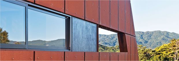 Close-up exterior view of a contemporary building facade clad in reddish-brown metal panels, with a large dark-framed window and a deep angular opening revealing mountainous terrain beyond. The facade has visible panel joints and exposed fasteners, creating a crisp, geometric architectural composition.