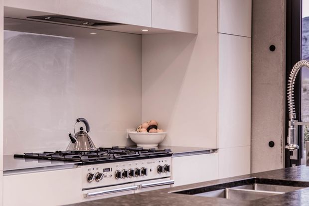 Close-up view of a refined contemporary kitchen featuring pale seamless cabinetry, a matching backsplash, an integrated rangehood, a stainless-steel freestanding gas range, dark stone benchtops, and a double-bowl undermount sink with a sculptural spring-coil tap. A small bowl of fruit sits on the cooktop area.