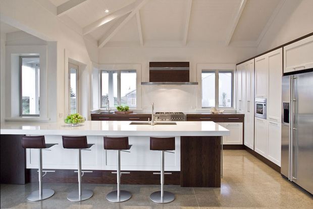 Bright, spacious contemporary kitchen with a large white island and four bar stools, dark timber cabinetry, white built-in wall cabinetry, stainless-steel appliances, and a polished light stone or concrete floor. The room features a high white pitched, exposed-beam ceiling and abundant windows.