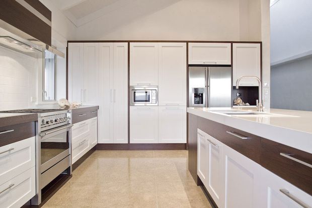 Bright, spacious contemporary kitchen with white Shaker-style cabinetry, dark timber-look drawer fronts, a large central island with an integrated sink, stainless-steel appliances, and pale tiled flooring. The layout includes extensive built-in storage, a freestanding oven, refrigerator, microwave, and a raised breakfast-bar area. The room has a clean, refined, high-contrast palette with warm neutral finishes.