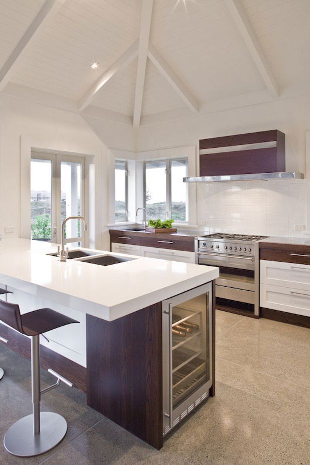 Bright, spacious contemporary kitchen with a large white island, integrated double sink, dark timber cabinetry, stainless-steel appliances, and bar seating. The kitchen has expansive windows and glazed doors, a polished concrete floor, white tiled splashback, and a high vaulted ceiling with exposed painted structural beams.