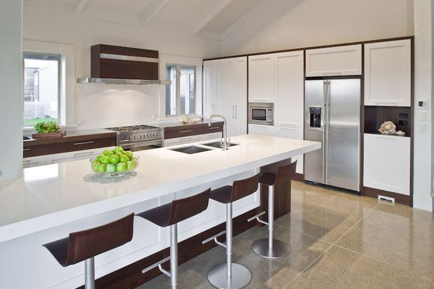 Bright, spacious contemporary kitchen with extensive white cabinetry, dark timber accents, stainless-steel appliances, a large white island with integrated sink and seating for four, and polished neutral-toned flooring. White coffered ceiling details and abundant glazing add an airy, refined feel.