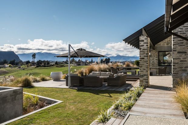 A contemporary mountain-side outdoor entertaining area beside a stone-clad building. The landscaped terrace includes timber decking, built-in concrete edging, outdoor lounge furniture, a large umbrella, ornamental grasses, and expansive views toward rugged mountains and open countryside. The building has dark metal roof elements, substantial grey stone walls, and large glazed openings.
