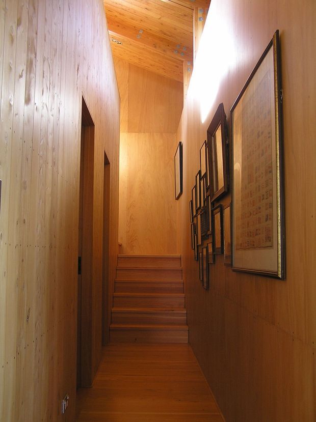 A narrow timber-lined hallway leading to a short flight of timber stairs. The walls, ceiling, stair treads and floor are clad in warm honey-toned wood, with framed artwork arranged along the right wall and doorways opening off the left side. The corridor has a tall, enclosed vertical form with bright natural light entering from above or ahead.