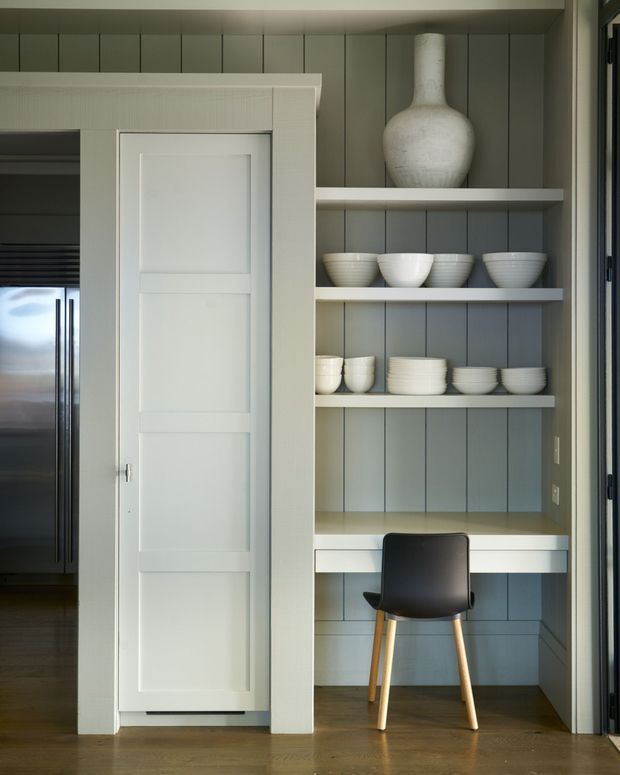 A compact built-in study nook beside a partially open paneled door, featuring grey vertical wall panelling, white open shelving displaying ceramic vessels, a white desk surface, and a simple black-and-timber chair. A stainless refrigerator is visible through the doorway, with timber flooring throughout.