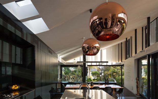 A sophisticated open-plan kitchen and dining space with a strong indoor-outdoor connection. The room features a long island with a reflective light stone benchtop, dark built-in cabinetry along the left wall, dramatic polished copper spherical pendant lights, black-framed glazing, skylights, and a large dining table overlooking a landscaped garden. The interior has a refined contemporary character with high ceilings and abundant natural light.