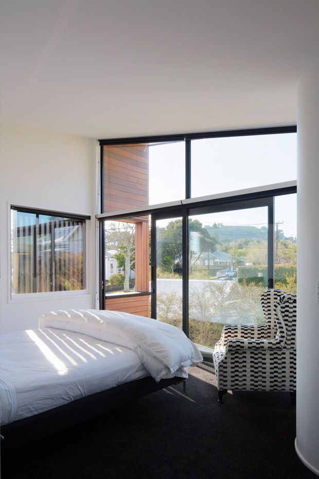 A bright contemporary bedroom with a low-profile bed dressed in white bedding, expansive black-framed floor-to-ceiling glazing, warm vertical timber wall cladding, and a bold black-and-white patterned armchair. Sunlight casts strong linear shadows across the bed, while a suburban landscape is visible outside.