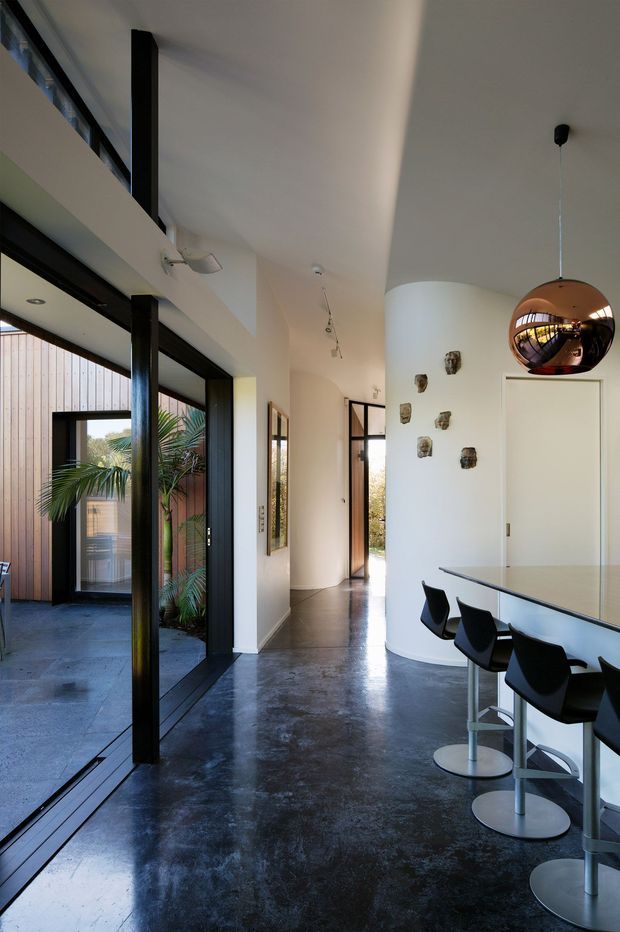 A contemporary open-plan interior combining a kitchen and circulation area. The space features a long white island with black bar stools, polished dark concrete flooring, expansive black-framed glazing, a curved white wall, and a reflective spherical pendant light. A planted outdoor courtyard and timber-clad exterior are visible through the glazing.