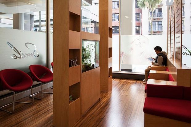 Bright contemporary commercial reception or waiting area with warm timber flooring and a series of tall timber room-divider joinery panels containing open display niches. Red upholstered lounge chairs and bench seating line the space, while full-height glazing, frosted graphics, and visible urban buildings create an airy office or public-facing interior. A seated person is present near the window.