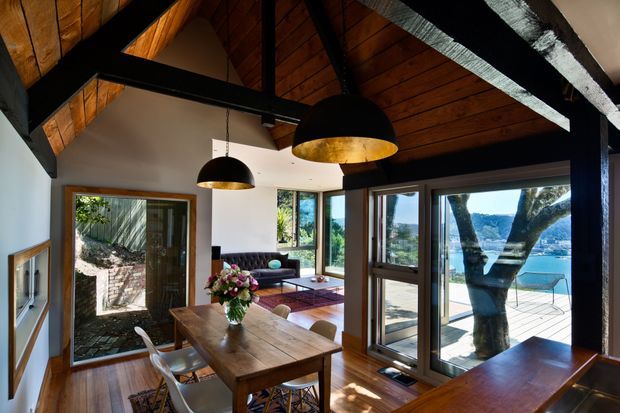 A warm, timber-lined dining and living area with dramatic exposed dark structural beams and a pitched wood ceiling. A solid timber dining table sits beneath two black pendant lights with gold interiors, while large glazed windows and doors frame a bright waterfront view and outdoor terrace. The interior includes timber flooring, white walls, a small seating area, and built-in timber surfaces, creating a rustic yet refined contemporary atmosphere.