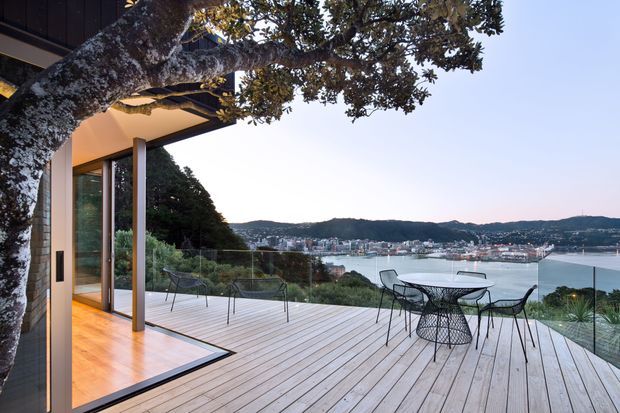 A modern home opening onto a spacious timber deck overlooking a sheltered bay and dense coastal settlement. The deck includes a round outdoor dining table with wire-frame chairs, while full-height glazing connects it to a warmly lit interior. A substantial tree frames the foreground and the house has a clean, flat-roofed architectural form with dark exterior cladding and slender glazed openings.