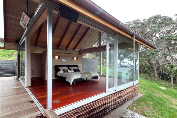 A compact, elevated bedroom pavilion with extensive floor-to-ceiling glass sliding walls opening onto a timber deck. The room features a pitched exposed-timber ceiling with dark structural rafters, polished reddish timber flooring, a concrete feature wall behind twin beds, and a timber-clad exterior base. The pavilion sits within a lush landscaped setting, with trees and a distant blue-water view visible beyond the glazing.