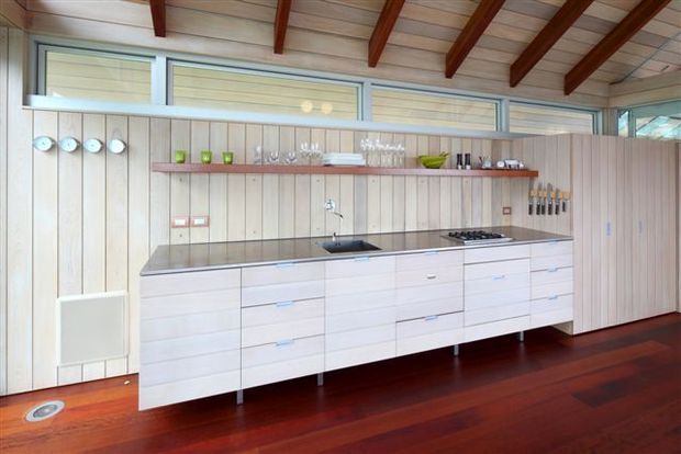 A bright, linear kitchen with extensive pale built-in cabinetry, a dark stone-look benchtop, integrated cooktop and sink, open shelving, and a full-height timber-panelled wall. High clerestory-style glazing and exposed reddish timber ceiling beams bring natural light and warmth, while the dark red-brown timber floor contrasts with the light joinery.