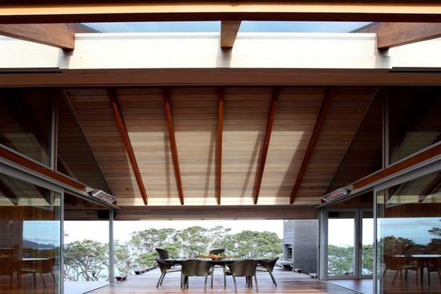 A contemporary open-plan dining space framed by extensive floor-to-ceiling glazing, with a timber-lined ceiling, exposed dark timber beams and rafters, and a central skylight. A dining table and chairs sit at the edge of the glazed opening, with trees and water visible beyond. The space has a strong indoor-outdoor connection and abundant natural light.
