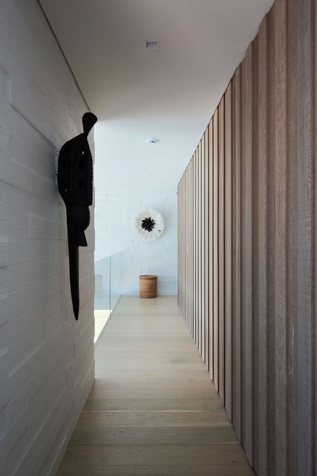 A narrow, minimalist interior hallway with pale timber flooring, a textured white wall on the left, and a full-height vertical timber slat feature wall on the right. A dark sculptural wall hanging, glass balustrade, small woven stool, and monochrome decorative artwork add restrained visual interest. The corridor is bright and calm, with clean contemporary detailing.