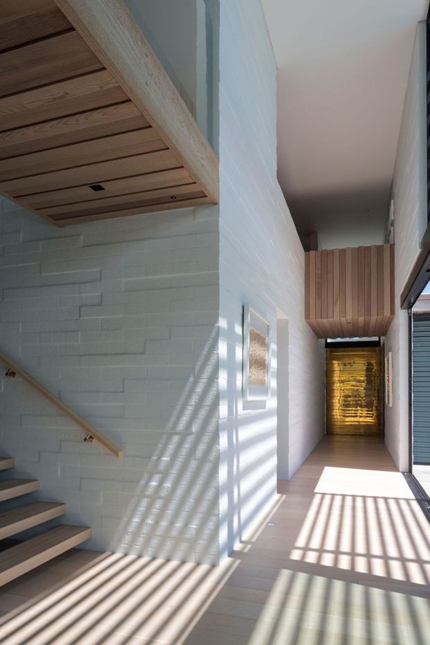 A bright, narrow modern hallway beside an open stairway. White-painted brick walls and pale timber flooring are illuminated by strong linear shadows from exterior screening or louvres. The ceiling and stair soffit feature exposed timber boards, while a timber-clad overhead volume and warm illuminated timber wall create a focal point at the far end. A glazed opening on the right connects the corridor to the exterior.