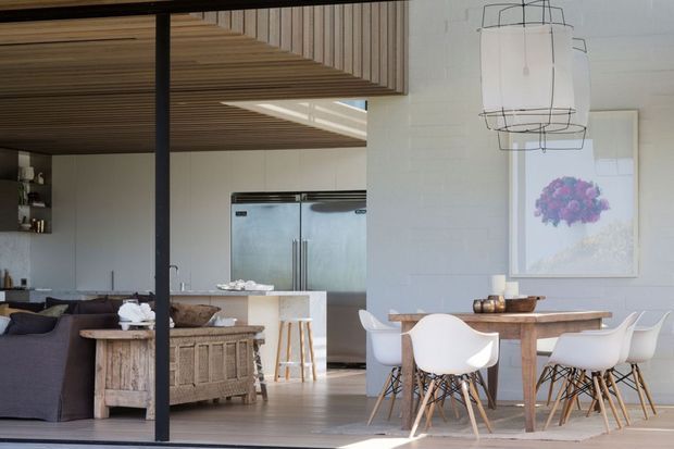 Open-plan contemporary interior showing a dining area in the foreground and a kitchen and living area beyond. The room features a timber slatted ceiling, pale painted brick walls, a dark framed glazed partition or opening, a white kitchen island with integrated stainless-steel refrigeration, a rustic timber dining table, white molded dining chairs, and a dark sofa. A large sculptural pendant light and framed artwork add decorative accents.