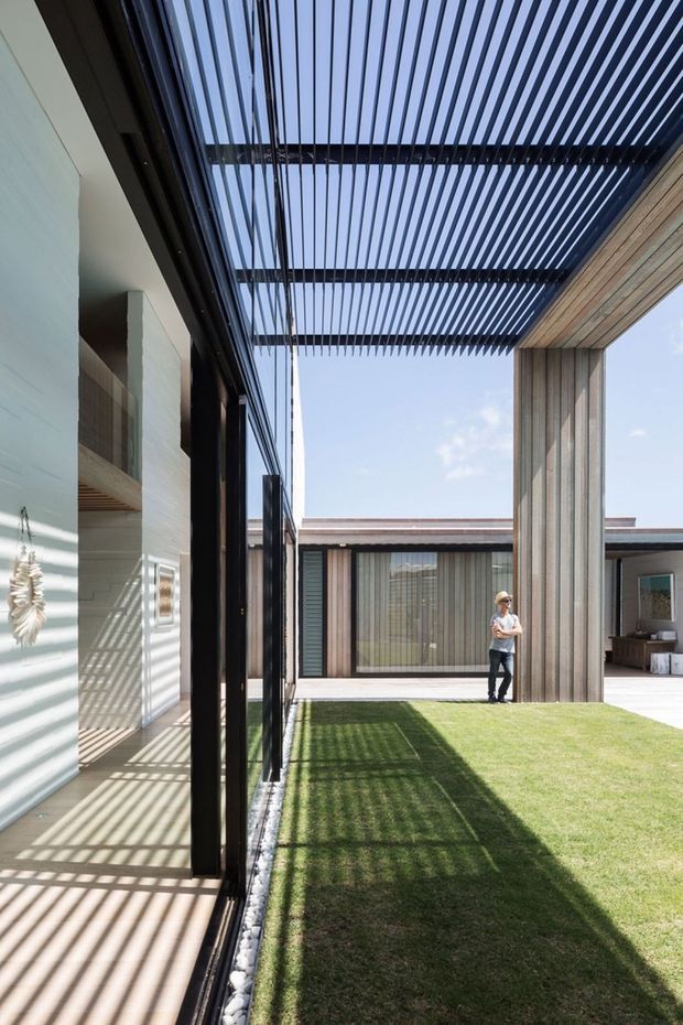 A contemporary courtyard framed by a modern residence, with extensive floor-to-ceiling glazing, vertical timber-clad walls, a broad black slatted pergola overhead, and a narrow lawn running through the outdoor space. Strong linear shadows fall across the paving and grass, creating a structured indoor-outdoor connection.
