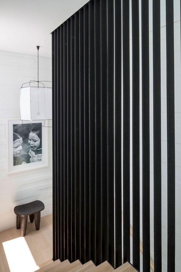 A minimalist interior stairway with pale timber steps beside a dramatic full-height black vertical slatted partition or balustrade. The adjoining white room features light timber flooring, a sculptural pendant light, framed monochrome artwork, and a small dark wooden stool. Strong natural light creates a high-contrast, refined atmosphere.